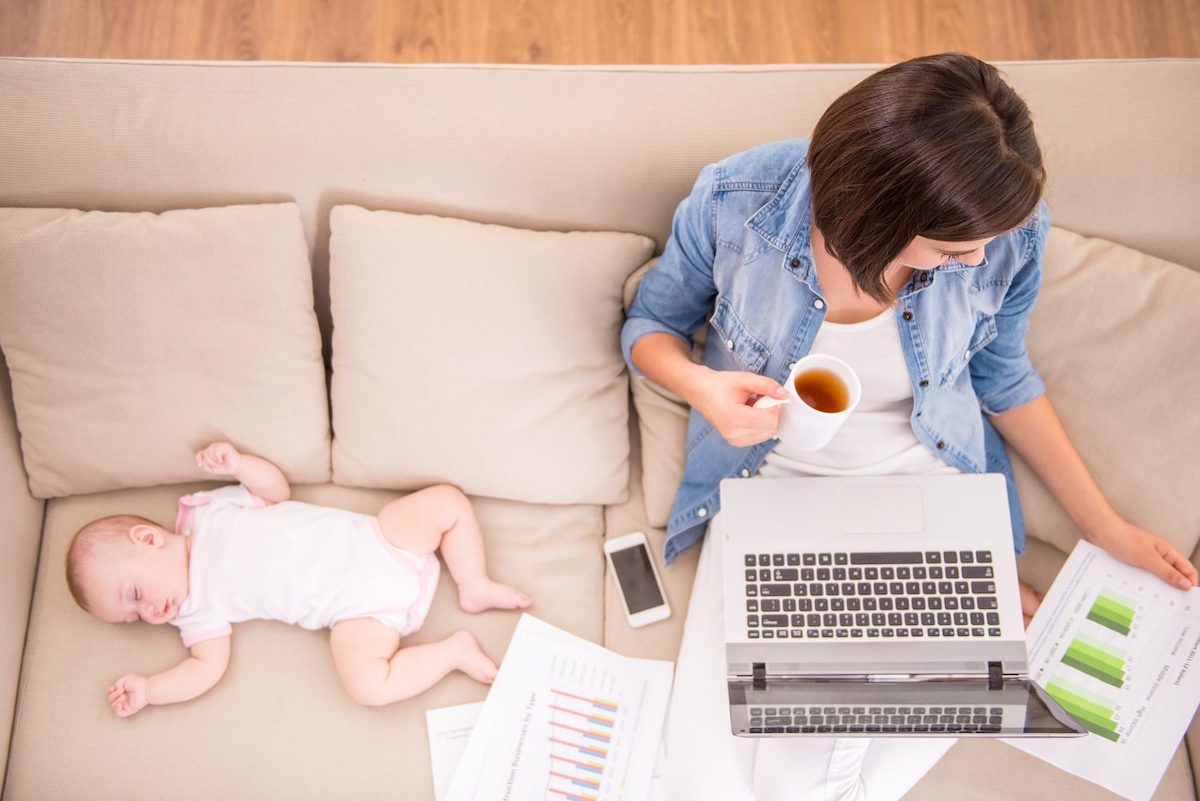 woman balancing work and life - siting on a couch with coffee and laptop with baby sleeping next to her